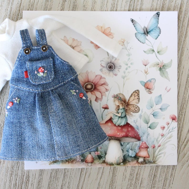 May include: A denim overall dress with embroidered mushroom details, paired with a white long-sleeved shirt. A floral and butterfly illustration is in the background, featuring a fairy sitting on a mushroom.