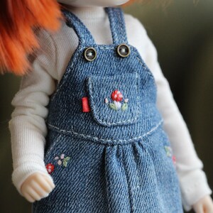 May include: A denim overall dress with embroidered mushroom and floral details. The dress features a small pocket and red accents. The doll is wearing a white long-sleeved shirt underneath the dress. The overall straps have decorative buttons.