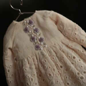 May include: A cream-coloured, crocheted dress with a floral embroidery design on the bodice. The dress has a round neckline and long sleeves.
