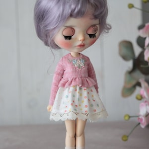 May include: A doll wearing a pink ruffled top with embroidered flowers and a white skirt with a floral print and lace trim.