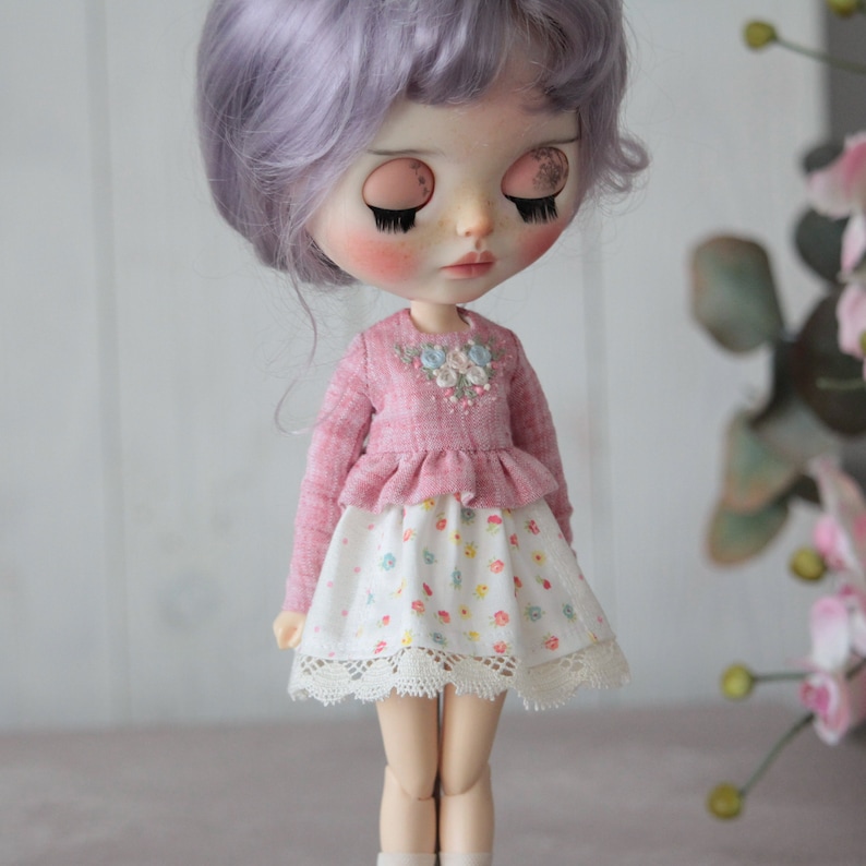 May include: A doll wearing a pink ruffled top with embroidered flowers and a white skirt with a floral print and lace trim.