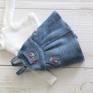 May include: A miniature denim dress with embroidered red flowers and a small pocket, paired with a white long-sleeved shirt. The dress features small metal buttons and delicate floral embroidery, creating a charming, handcrafted look.