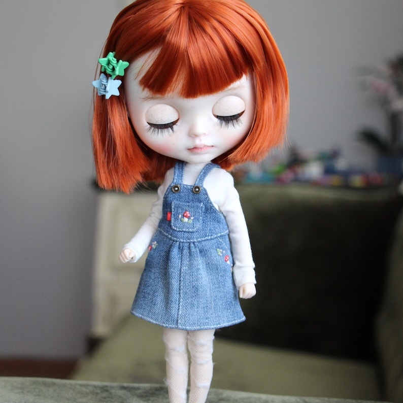 May include: A doll with red hair and closed eyes, wearing a denim overall dress over a white long-sleeved shirt. The dress features a small pocket with a mushroom design and floral embroidery. The doll has two small star-shaped hair clips.