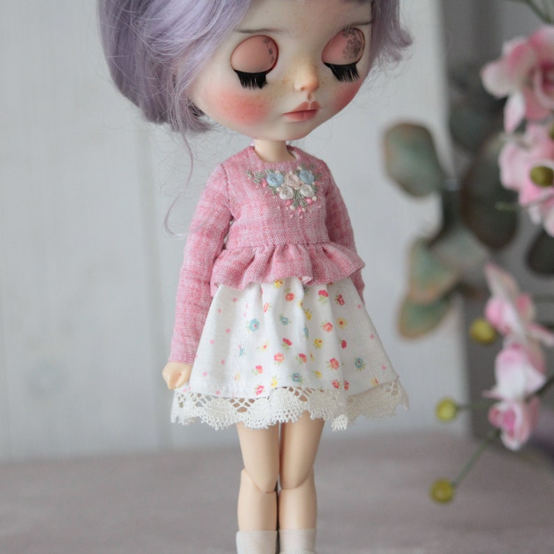 May include: A pink and white doll wearing a pink sweater with embroidered flowers and a white skirt with a floral print and lace trim.
