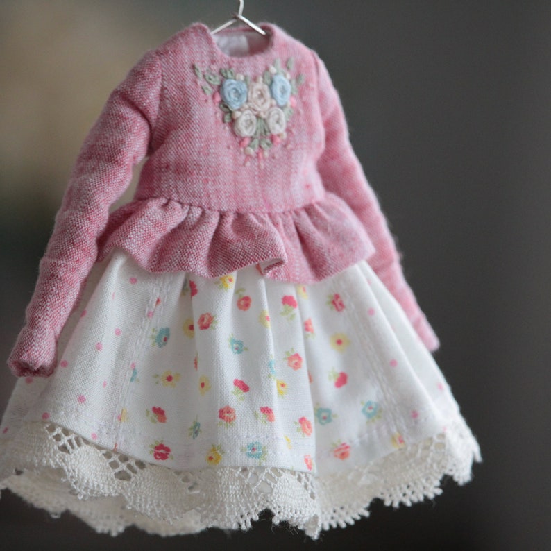 May include: A pink and white doll's dress with a floral embroidered bodice and a white skirt with a floral print and lace trim.