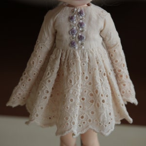 May include: A cream-coloured doll's dress with a delicate lace pattern and a floral embroidery design on the bodice.