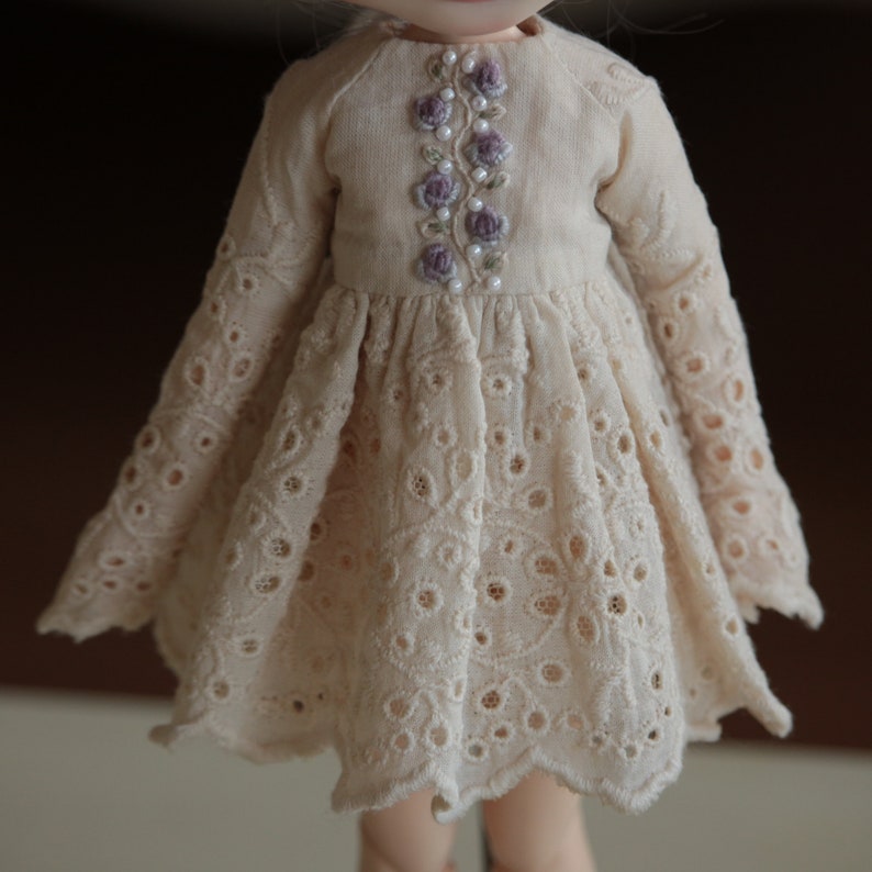 May include: A cream-coloured doll's dress with a delicate lace pattern and a floral embroidery design on the bodice.