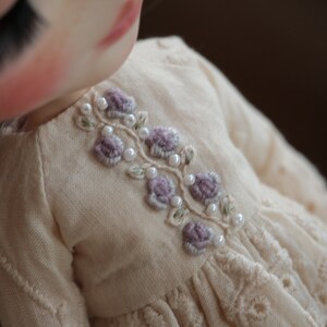 May include: A close-up of a cream-coloured dress with a delicate floral embroidery design. The embroidery features small purple flowers and white pearl beads.