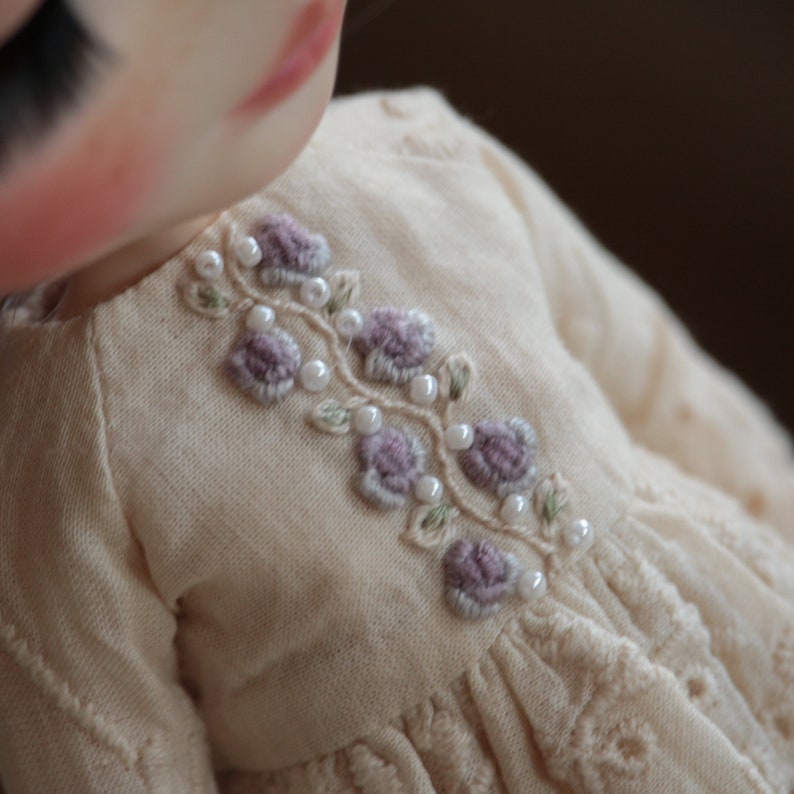 May include: A close-up of a cream-coloured dress with a delicate floral embroidery design. The embroidery features small purple flowers and white pearl beads.