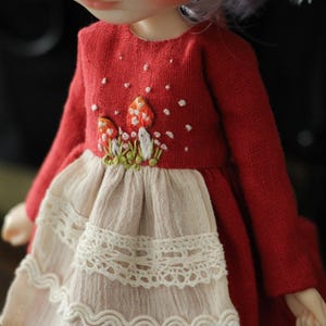 May include: A red dress with long sleeves and a white lace trim. The dress has a mushroom embroidery design on the front.