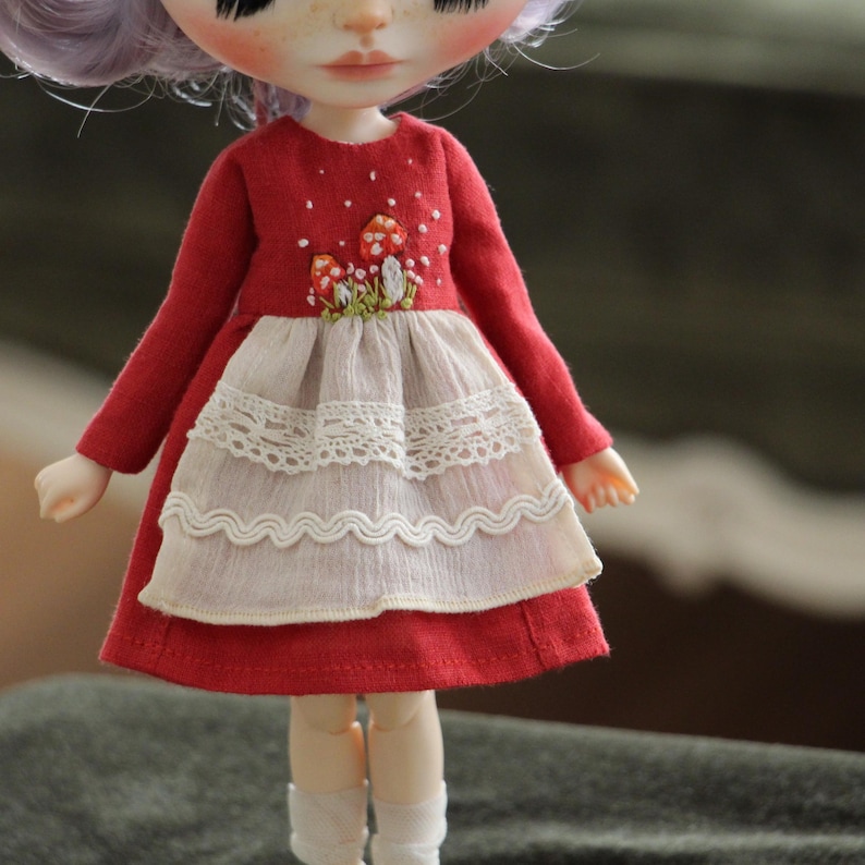 May include: A red dress with long sleeves and a white lace trim. The dress has an embroidered design of mushrooms and flowers on the front.
