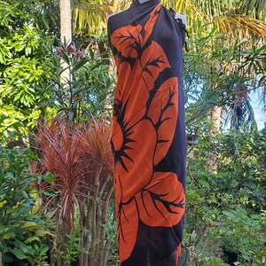 Puede incluir: Un sarong negro con un gran estampado de flor de hibisco naranja. El sarong est&aacute; drapeado sobre un maniqu&iacute; y se muestra sobre un fondo de exuberante follaje verde.