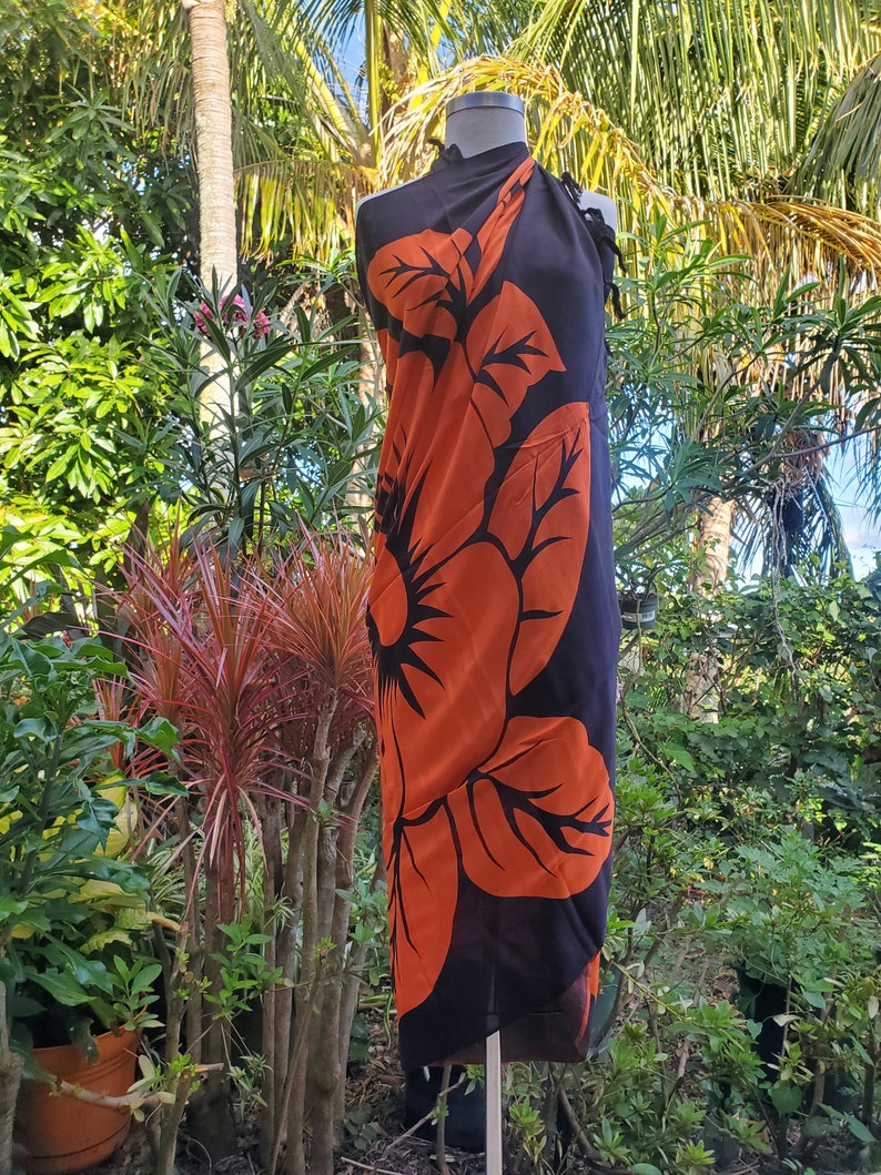 Puede incluir: Un sarong negro con un gran estampado de flor de hibisco naranja. El sarong est&aacute; drapeado sobre un maniqu&iacute; y se muestra sobre un fondo de exuberante follaje verde.