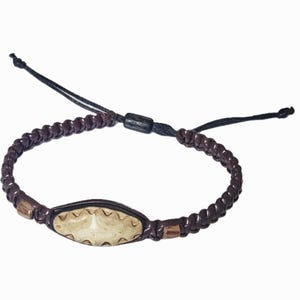 May include: Brown braided bracelet with a central oval-shaped beige accent piece. The accent has a carved design. The bracelet has a sliding knot closure with black cords. Two small wooden beads are on either side of the accent piece.