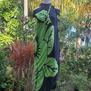 Puede incluir: Un sarong negro con un gran estampado floral verde. El sarong est&aacute; drapeado sobre un maniqu&iacute; y se exhibe al aire libre.