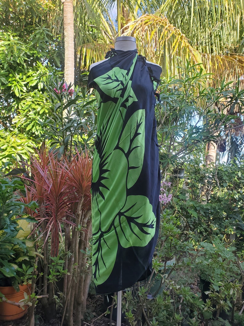 Puede incluir: Un sarong negro con un gran estampado floral verde. El sarong est&aacute; drapeado sobre un maniqu&iacute; y se exhibe al aire libre.