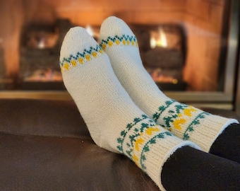May include: A pair of white knitted socks with yellow and green patterns. The socks are worn by a person sitting in front of a fireplace.