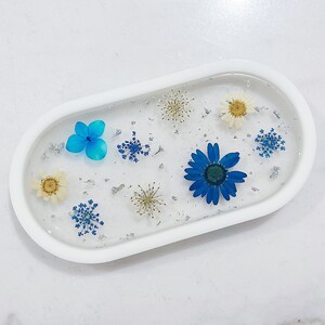 Peut inclure: Plateau ovale blanc avec un dessus en résine transparente rempli de fleurs pressées. Les fleurs sont bleues, blanches et jaunes. Le plateau est parfait pour présenter de petits objets ou des bijoux.