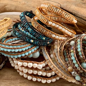May include: An assortment of wrap bracelets featuring various designs and materials. The bracelets include beads in shades of blue, white, and gold, as well as pearls and crystals. The bracelets are displayed on a piece of driftwood.