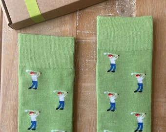 May include: Two pairs of light green socks with a repeating pattern of a golfer in a red hat, white shirt, and blue pants. The socks are displayed on a wooden surface, with a brown box and green ribbon in the background.