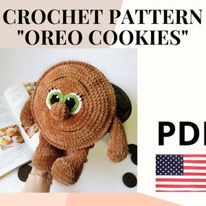 Puede incluir: Patrón de crochet para un juguete de peluche Oreo. El patrón está disponible para descargar en PDF. La imagen muestra un juguete de peluche marrón con ojos verdes y una cara sonriente. El juguete está sentado en un libro y un plato con galletas Oreo.