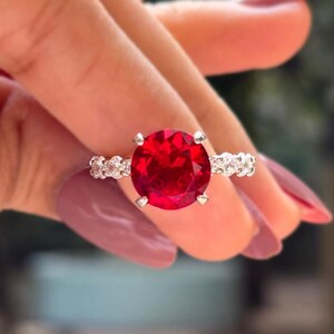 May include: A silver ring featuring a large, round, vibrant red gemstone as the centerpiece. The band is adorned with smaller, clear gemstones. The ring is held up against a blurred background, showcasing the ring's brilliance.