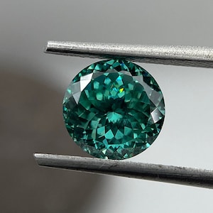 May include: A round, faceted, green gemstone with a slight yellow hue, held by tweezers against a white background.