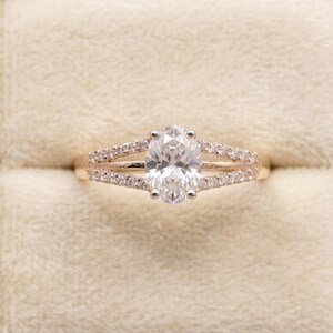 Op de afbeelding: Een gouden ring met een grote ovaal geslepen diamant als middensteen. De band splitst zich in twee rijen kleinere diamanten aan weerszijden, gepresenteerd in een fluwelen ringdoos. Klassiek ontwerp.