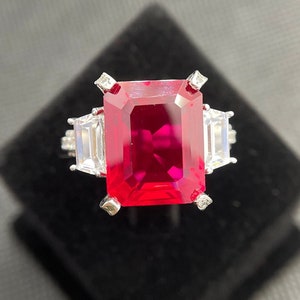 Puede incluir: Un anillo de plata con una gran piedra preciosa de rubí de talla esmeralda rodeada de piedras preciosas más pequeñas y transparentes.