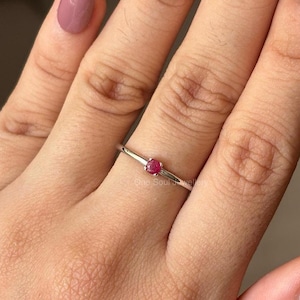 May include: A delicate silver ring with a small, round, red gemstone. The ring is worn on a finger with light pink nail polish.