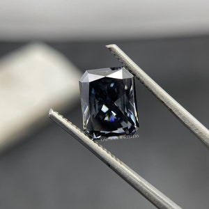 May include: A dark grey emerald cut gemstone held by tweezers. The gemstone has a slight blue hue and is faceted.