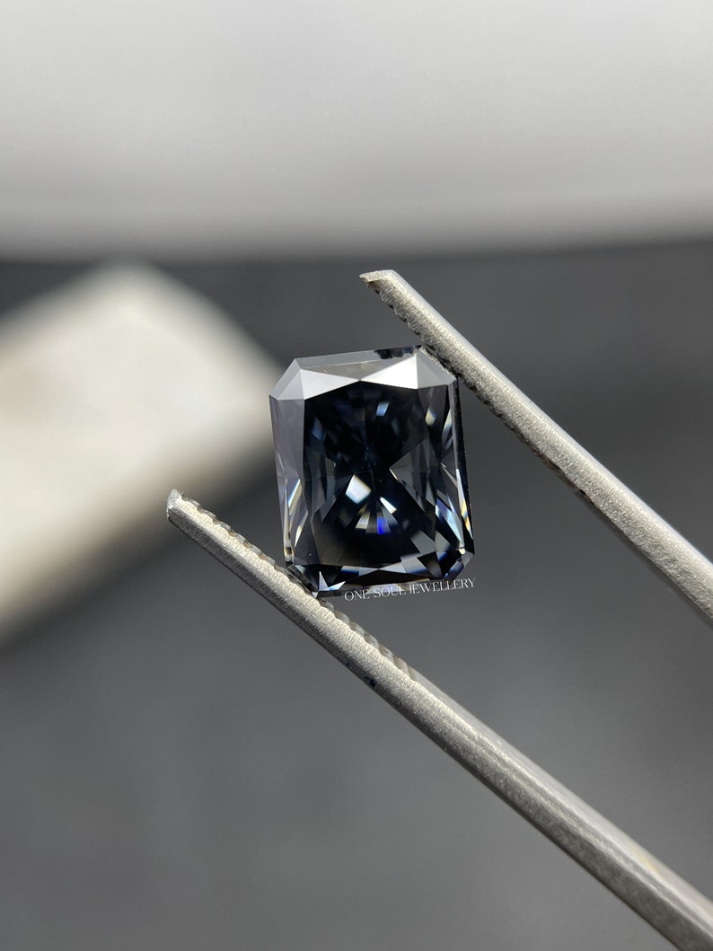 May include: A dark grey emerald cut gemstone held by tweezers. The gemstone has a slight blue hue and is faceted.