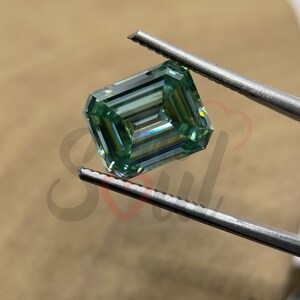 May include: A green emerald cut moissanite gemstone held by tweezers. The gemstone has a brilliant, faceted cut and a light green colour.