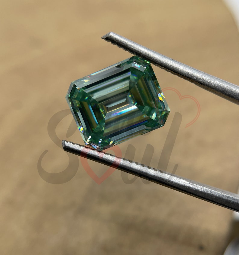 May include: A green emerald cut moissanite gemstone held by tweezers. The gemstone has a brilliant, faceted cut and a light green colour.