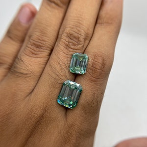 May include: Two emerald cut green gemstones, one larger than the other, are displayed on a hand.