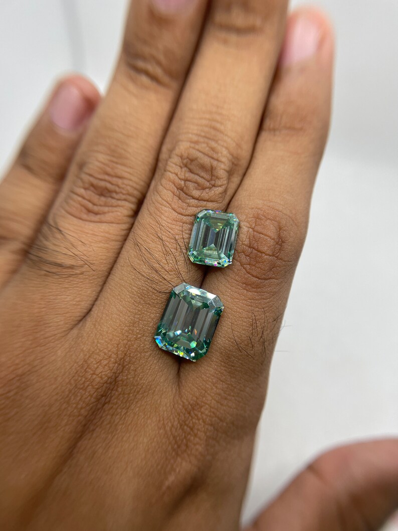 May include: Two emerald cut green gemstones, one larger than the other, are displayed on a hand.