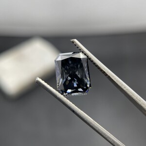 May include: A dark grey emerald cut gemstone held in tweezers. The gemstone has a slight blue hue and is set against a white background.