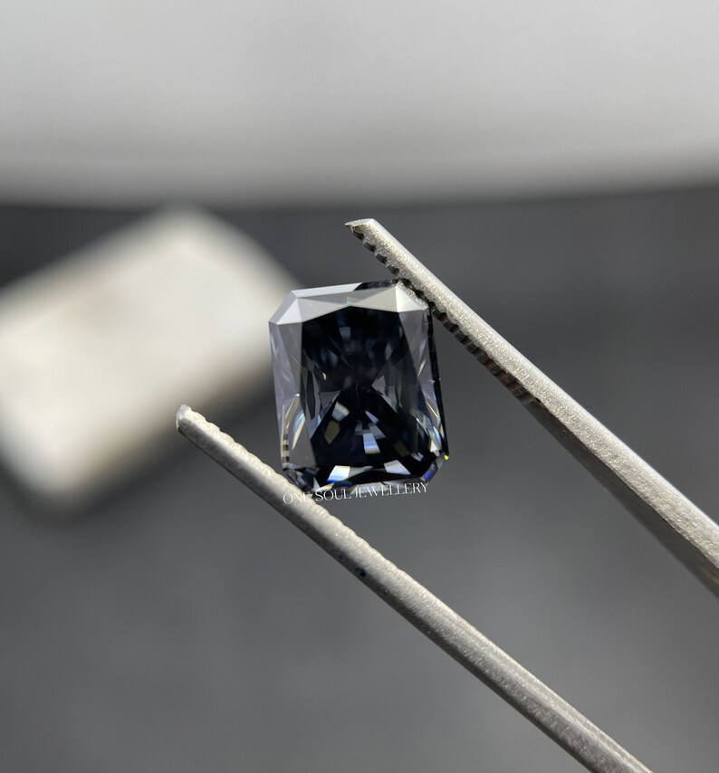 May include: A dark grey emerald cut gemstone held in tweezers. The gemstone has a slight blue hue and is set against a white background.