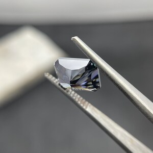 May include: A dark grey gemstone with a square shape and a faceted cut. The gemstone is held in tweezers.