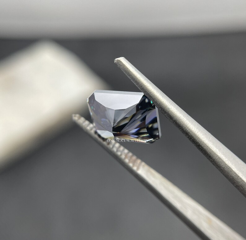 May include: A dark grey gemstone with a square shape and a faceted cut. The gemstone is held in tweezers.