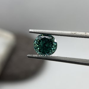 May include: A round, green gemstone with a faceted cut, held by tweezers. The gemstone has a deep green colour with a slight blue hue.  "ONE SOUL JEWELLERY" is printed on the tweezers.