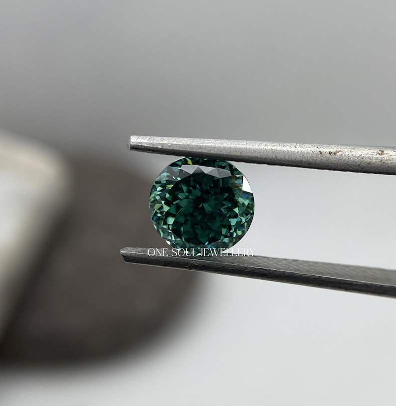 May include: A round, green gemstone with a faceted cut, held by tweezers. The gemstone has a deep green colour with a slight blue hue.  "ONE SOUL JEWELLERY" is printed on the tweezers.