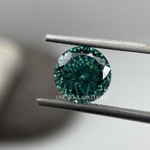May include: A round, faceted, green gemstone with a blue hue is held by tweezers. The gemstone is a vibrant green with a slight blue tint. The gemstone is held against a white background.