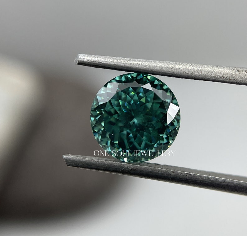 May include: A round, faceted, green gemstone with a blue hue is held by tweezers. The gemstone is a vibrant green with a slight blue tint. The gemstone is held against a white background.