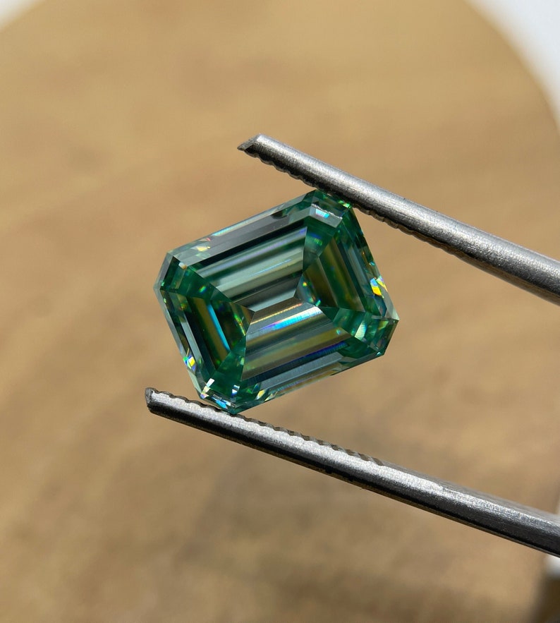 May include: A green emerald cut gemstone with a rainbow effect, held by tweezers.