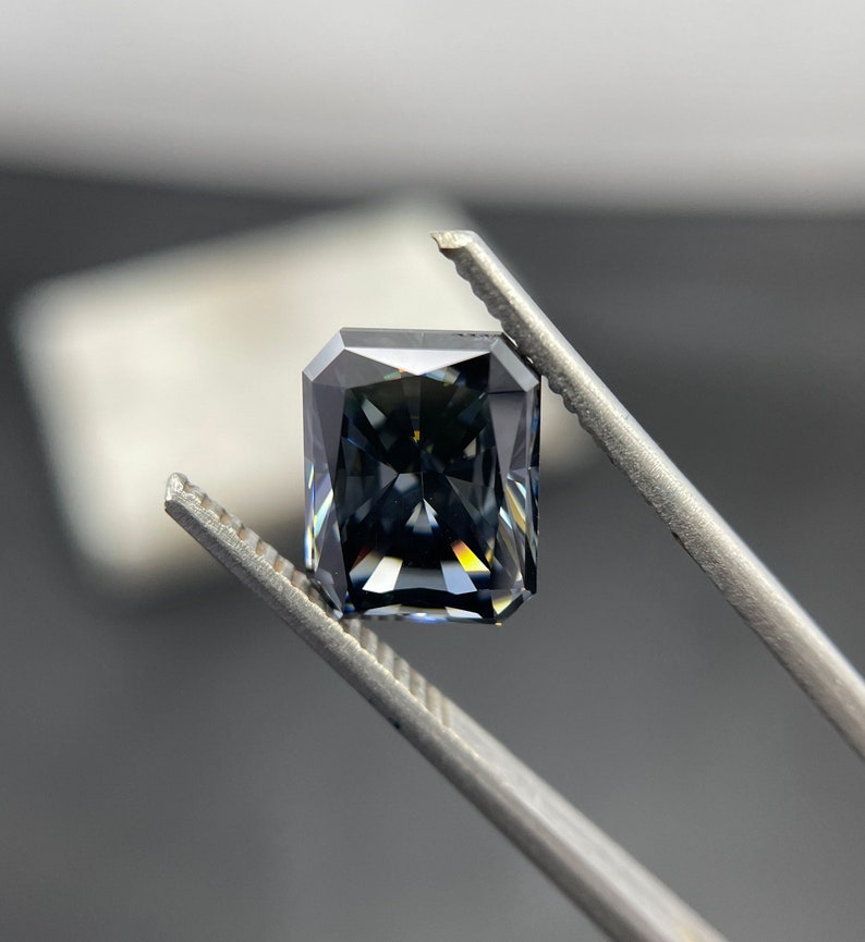 May include: A dark grey emerald cut gemstone held by tweezers. The gemstone has a slight blue hue and is faceted.