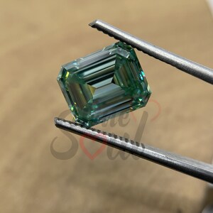 May include: A green emerald cut moissanite gemstone held by tweezers. The gemstone has a light green colour and a high level of brilliance.