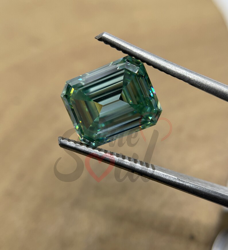 May include: A green emerald cut moissanite gemstone held by tweezers. The gemstone has a light green colour and a high level of brilliance.