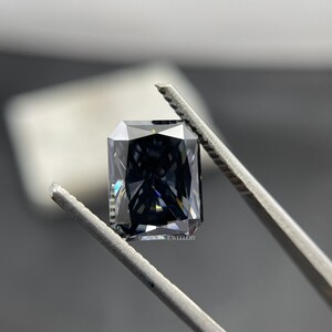 May include: A dark grey, square-shaped gemstone with a faceted cut, held by tweezers. The gemstone has a slight blue hue and reflects light.