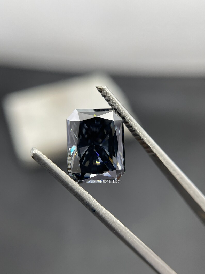 May include: A dark grey, square-shaped gemstone with a faceted cut, held by tweezers. The gemstone has a slight blue hue and reflects light.